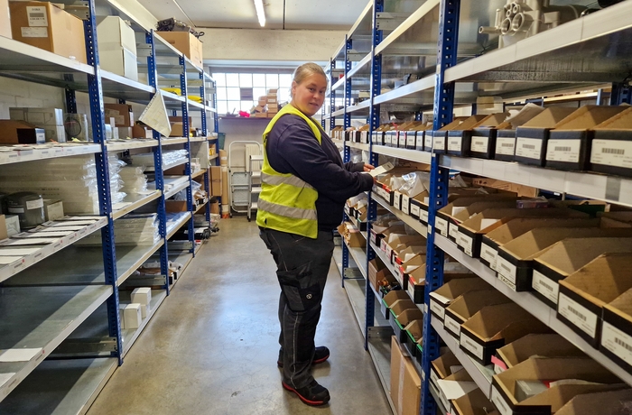 Sara Verstraeten met een fluohesje in het magazijn, tussen de rekken met dozen met onderdelen