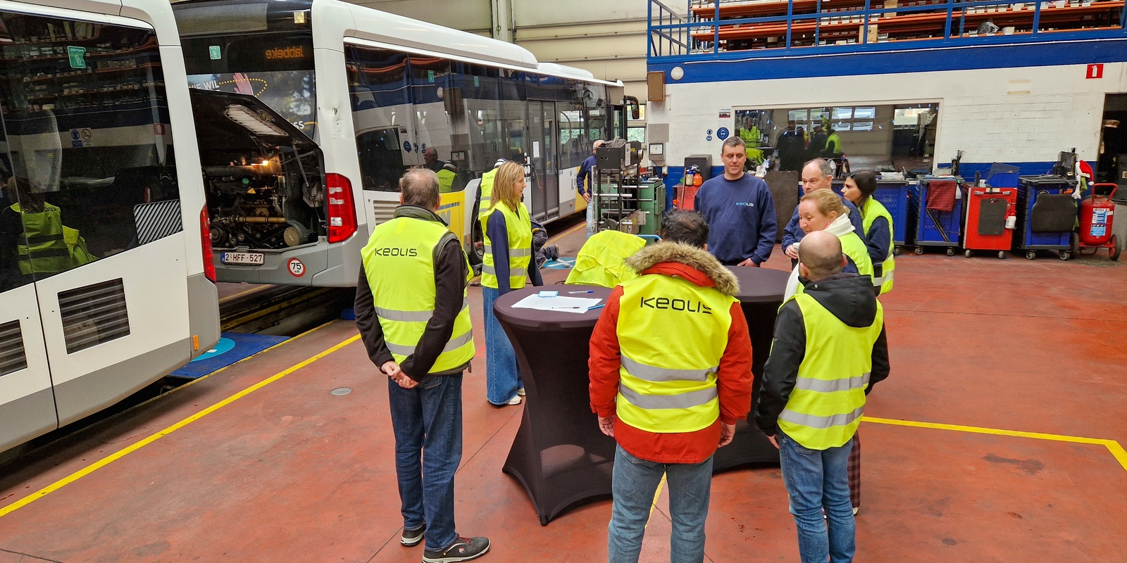 Groepje kandidaten in fluohesje staat aan een hoge tafel in de werkplaats.