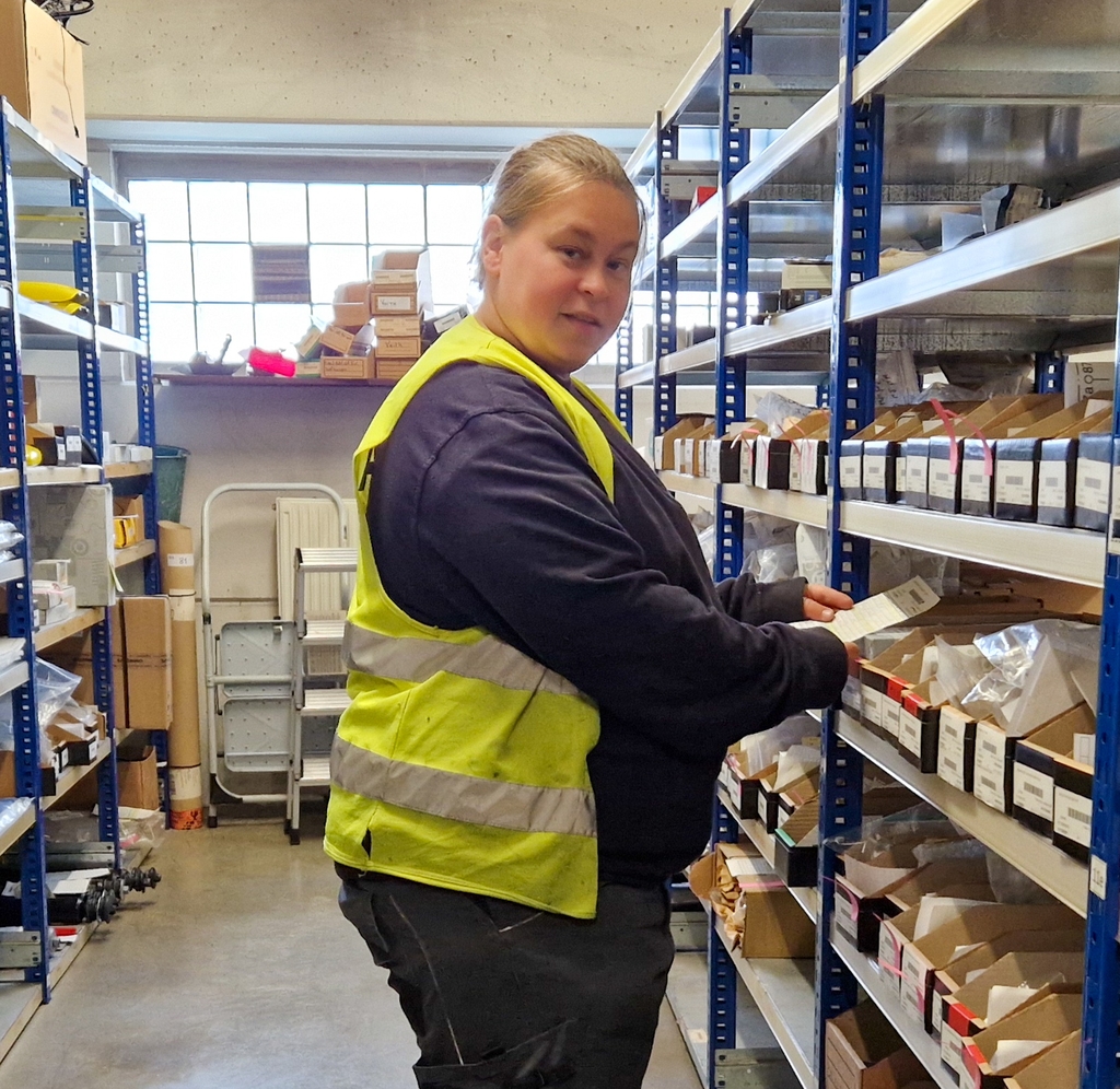 Sara Verstraeten met een fluohesje in het magazijn, tussen de rekken met dozen met onderdelen